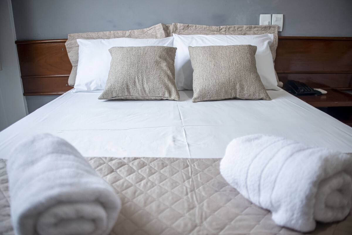 A neatly made bed with two beige pillows, a white pillow, and two rolled white towels on the bed.
