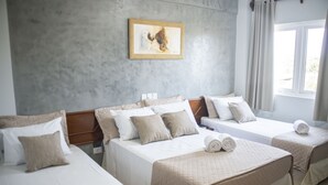 Quarto de hotel com três camas, cada uma com colcha acolchoada e travesseiros. Há uma pintura na parede acima das camas e uma janela com cortinas à direita.
