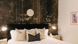 A bed with white bedding, a black headboard, and multiple pillows. There are two bedside tables with lamps. The room has a rustic wooden ceiling and a distressed wall.