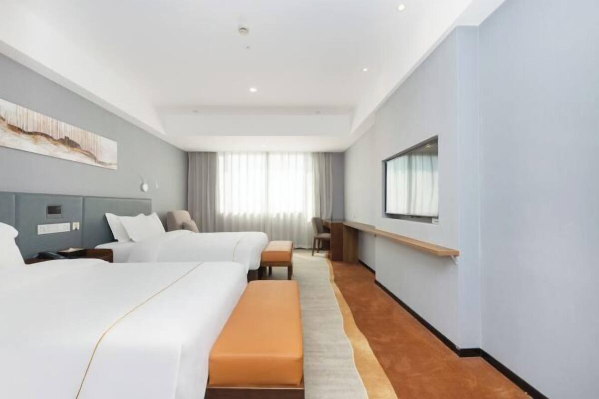 A hotel room with two beds, a desk, a chair, a large window with curtains, and a mounted TV.