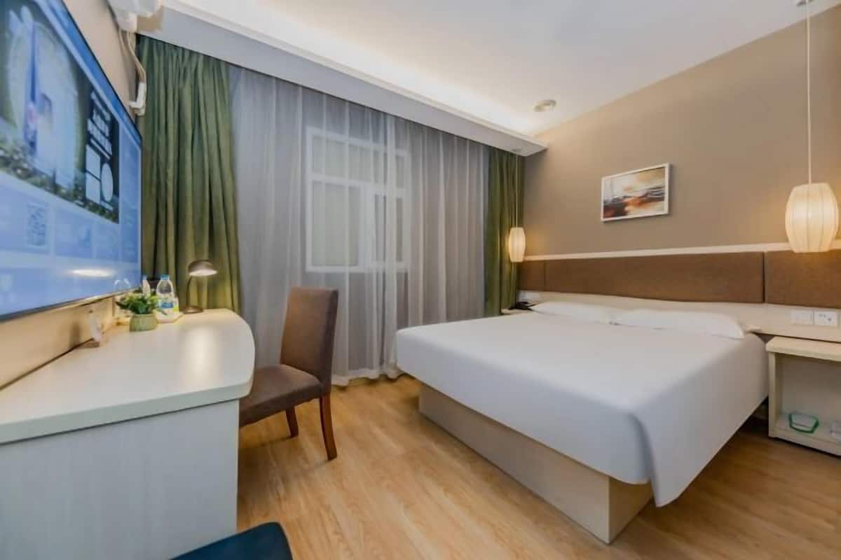 A hotel room with a bed, a chair, a desk with a lamp, a picture on the wall, and a window with curtains.