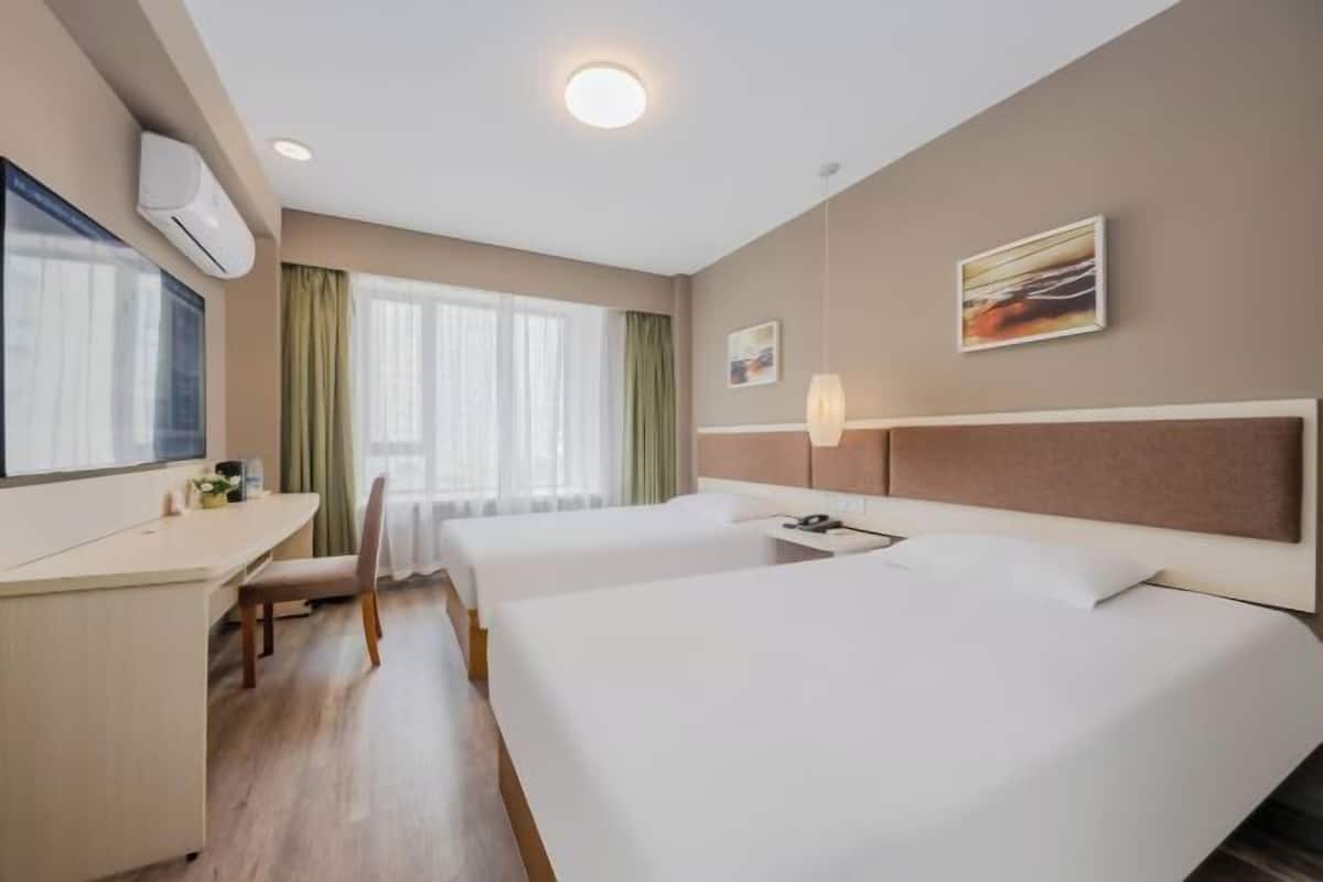 A hotel room with two beds, a desk, a chair, a window with curtains, and a wall-mounted air conditioner.