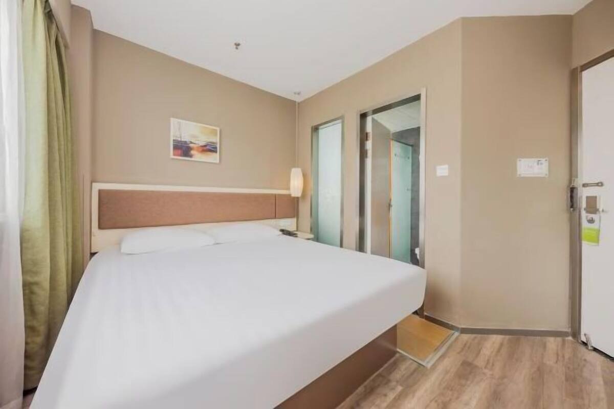 A hotel room with a bed, a painting on the wall, a door, and a light fixture.