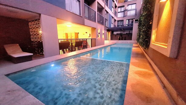 A swimming pool with lounge chairs, a covered seating area, and a building with balconies and a staircase. - Apto moderno com miniquintal e piscina (Rio de Janeiro)