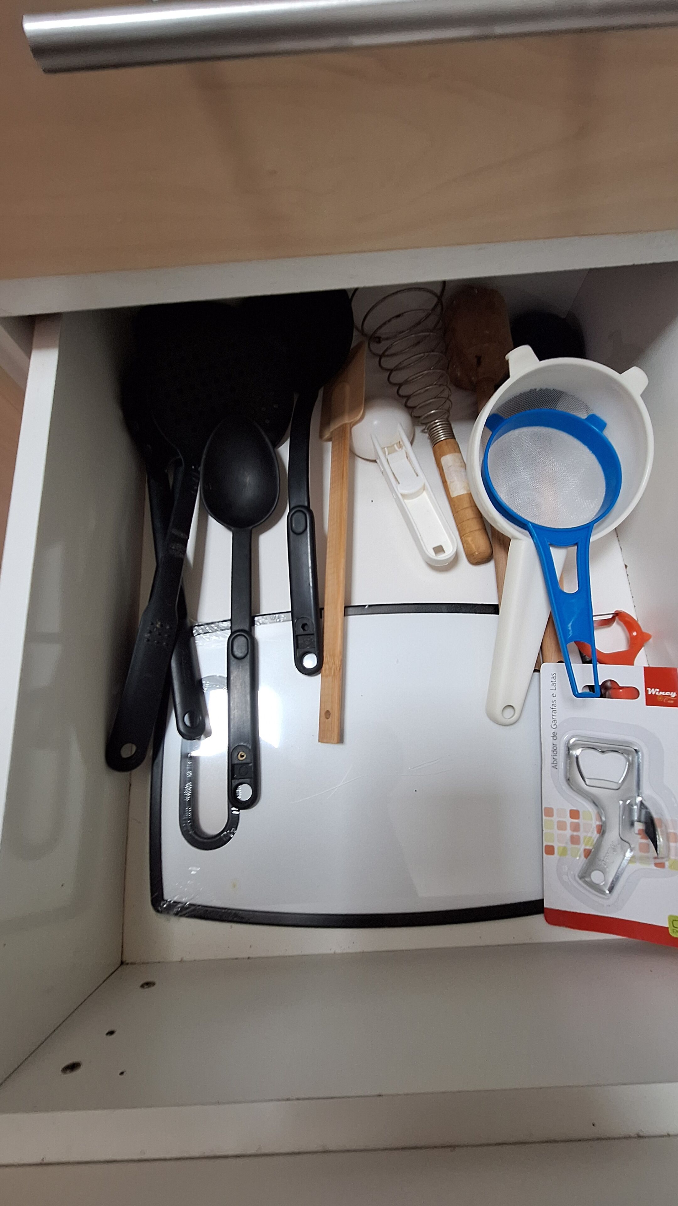 Un cajón de cocina con varios utensilios, incluyendo una cuchara grande negra, una espátula de madera y un colador blanco con mango azul.