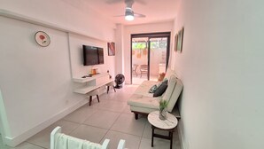 A living room with a sofa, a TV, a fan, and a small table with a plant. - Apto moderno com miniquintal e piscina (Rio de Janeiro)