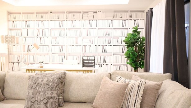 Una sala de estar con un sofá color beige, cojines estampados, una estantería llena de libros, una mesita con una lámpara y una planta verde.