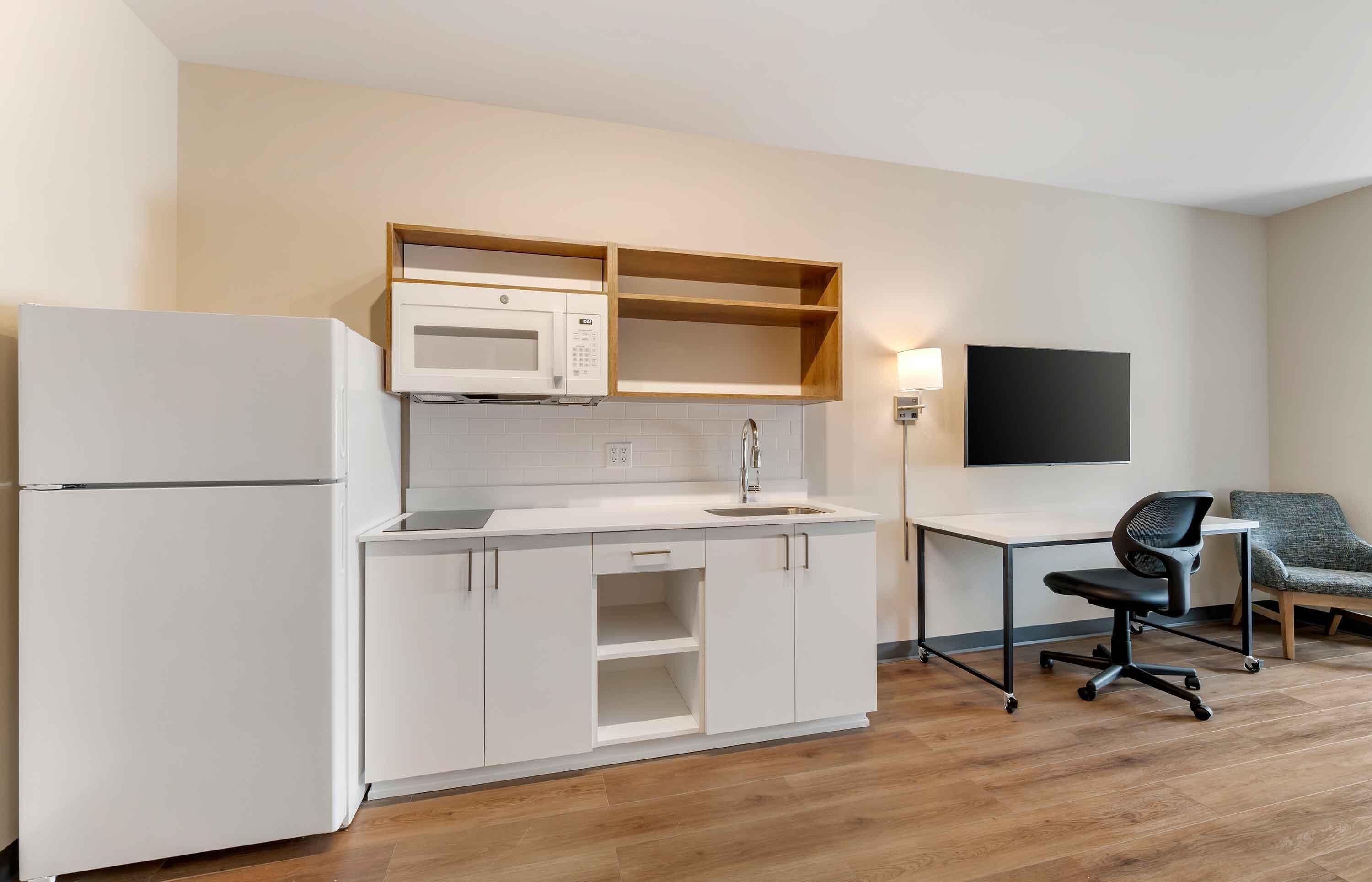 Una cocina compacta con refrigerador, microondas y lavamanos, una pequeña mesa de comedor con una silla, y un televisor de pantalla plana montado en la pared.