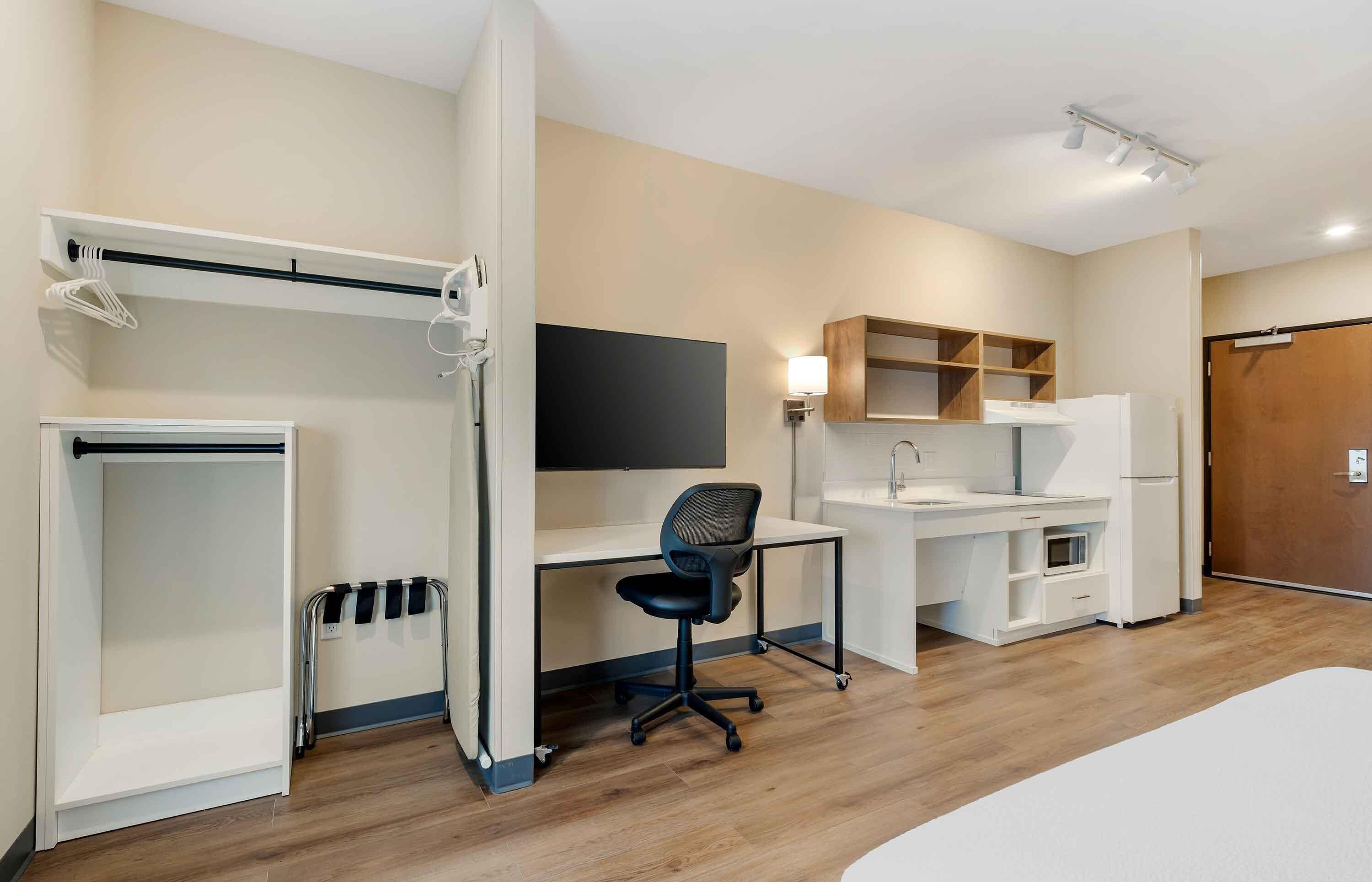A modern hotel room with a bed, a desk with a chair, a wardrobe, a TV, and a small kitchenette.