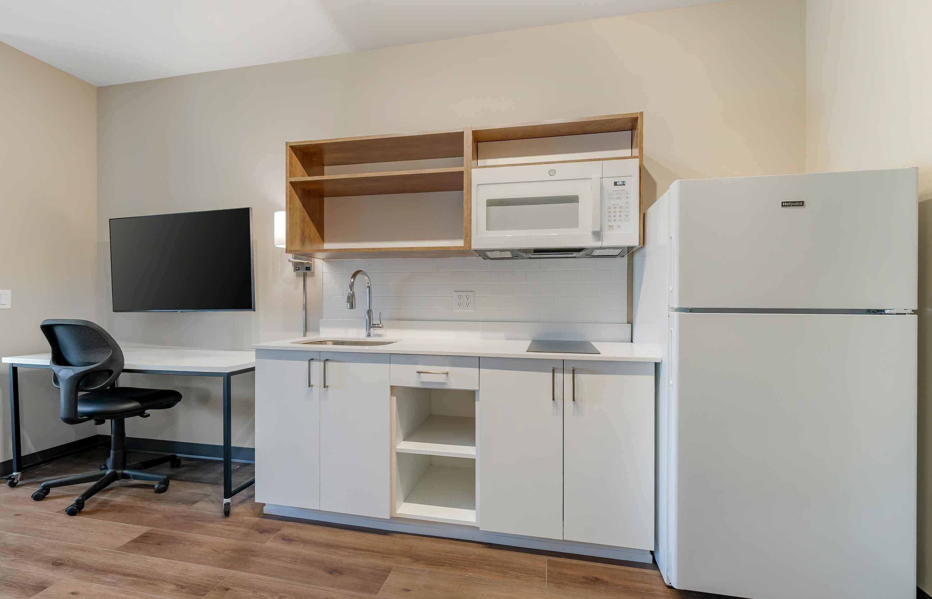Cocina compacta con microondas, refrigerador y un lavamanos pequeño. Hay un estante de madera sobre el microondas y una silla de oficina negra a la izquierda.