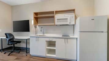 A compact kitchen with a microwave, refrigerator, and a small sink. There is a wooden shelf above the microwave and a black office chair to the left.