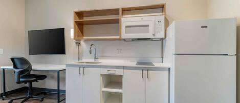 Cocina compacta con microondas, refrigerador y un lavamanos pequeño. Hay un estante de madera sobre el microondas y una silla de oficina negra a la izquierda.