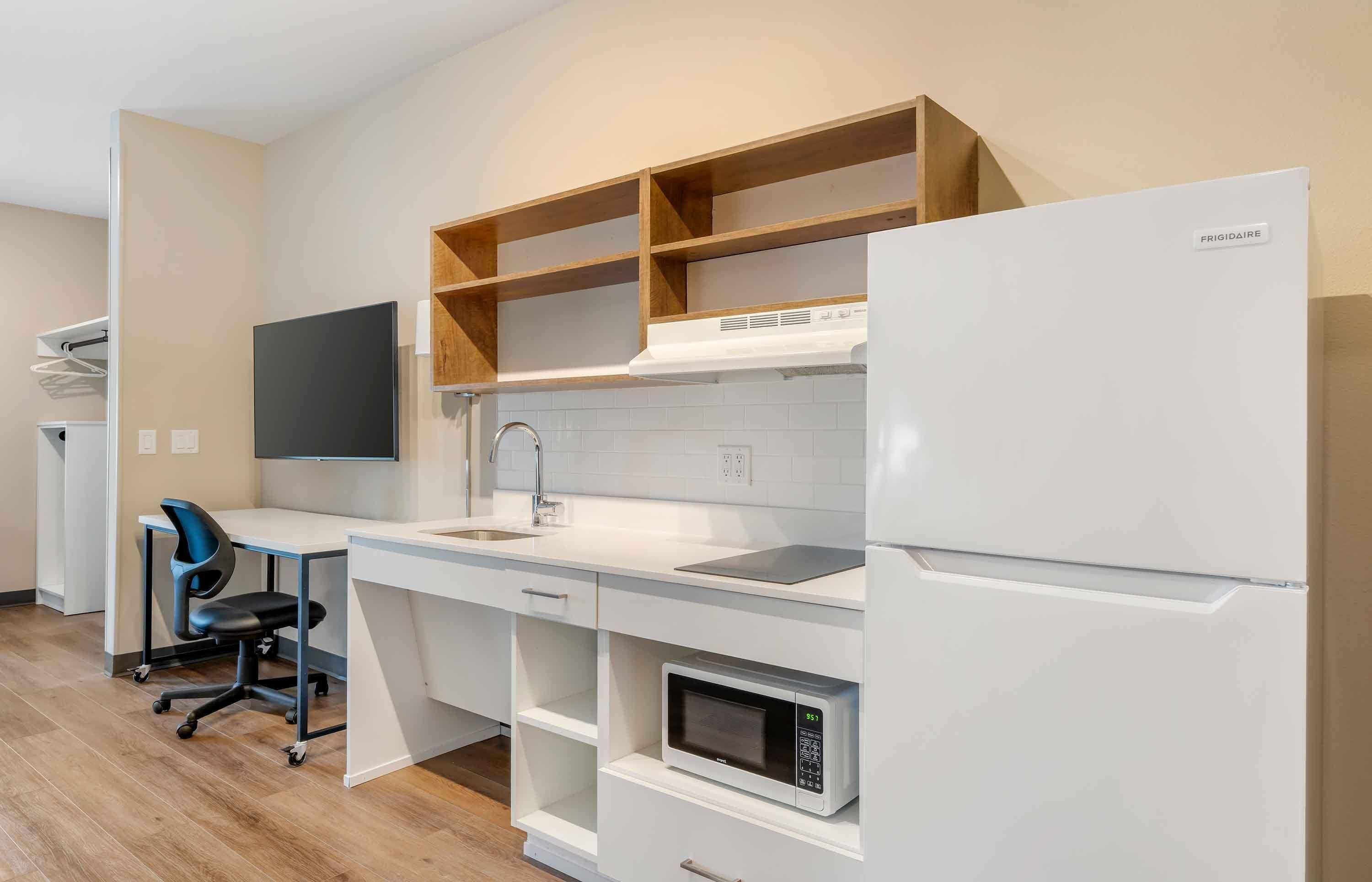 Una cocina compacta con un refrigerador blanco, un microondas, un lavamanos y una estantería de madera.