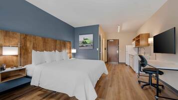 A hotel room with a bed, bedside table, lamp, desk with chair, and a painting on the wall.