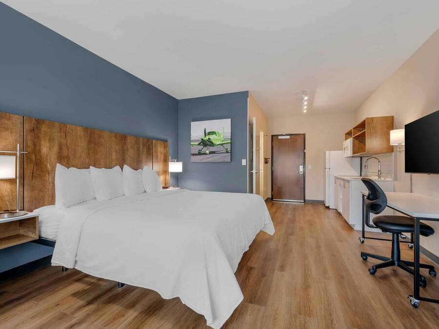 A hotel room with a bed, bedside table, lamp, desk with chair, and a painting on the wall.