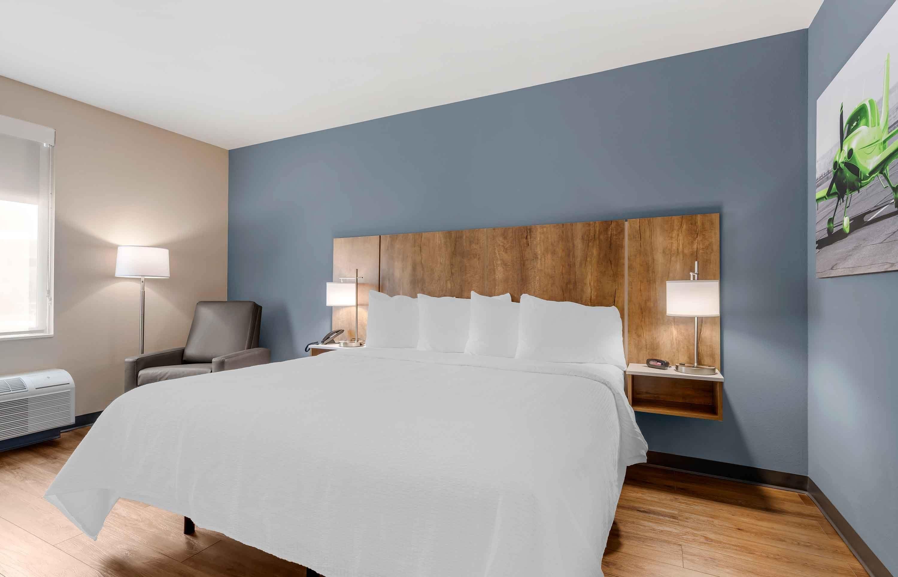 A hotel room with a large bed, a wooden headboard, two bedside tables with lamps, a chair, and a window with an air conditioner.