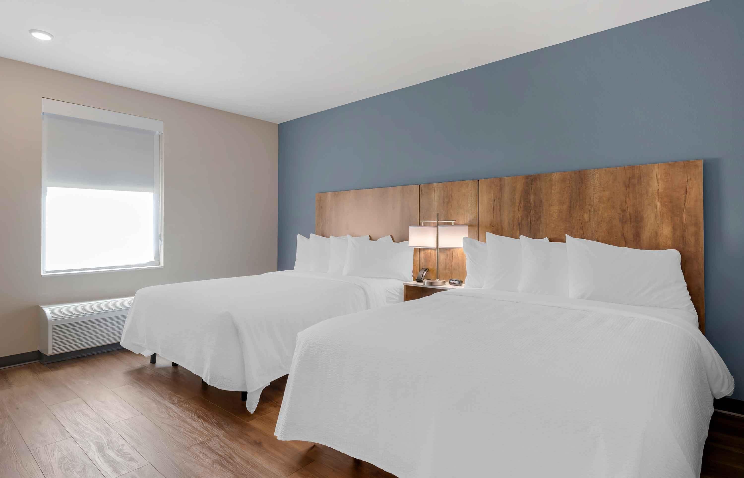A hotel room with two beds, a wooden headboard, a bedside table with a lamp, and a large window providing natural light.