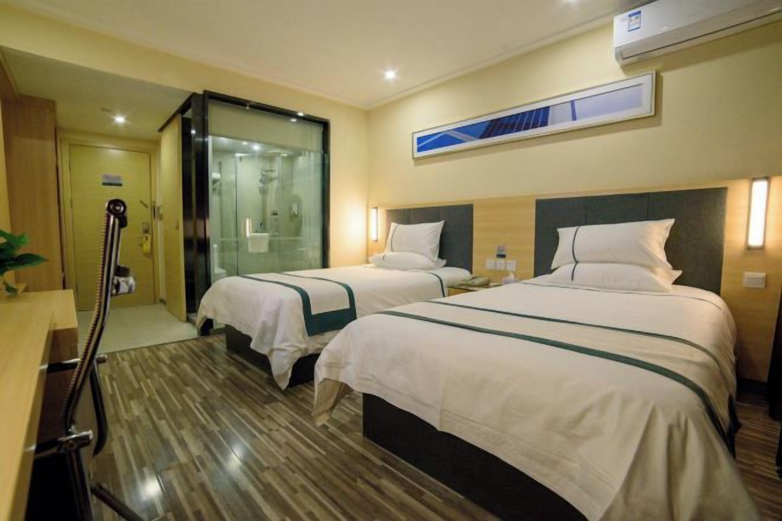 A hotel room with two beds, a wooden floor, a glass shower enclosure, and a mounted air conditioner.