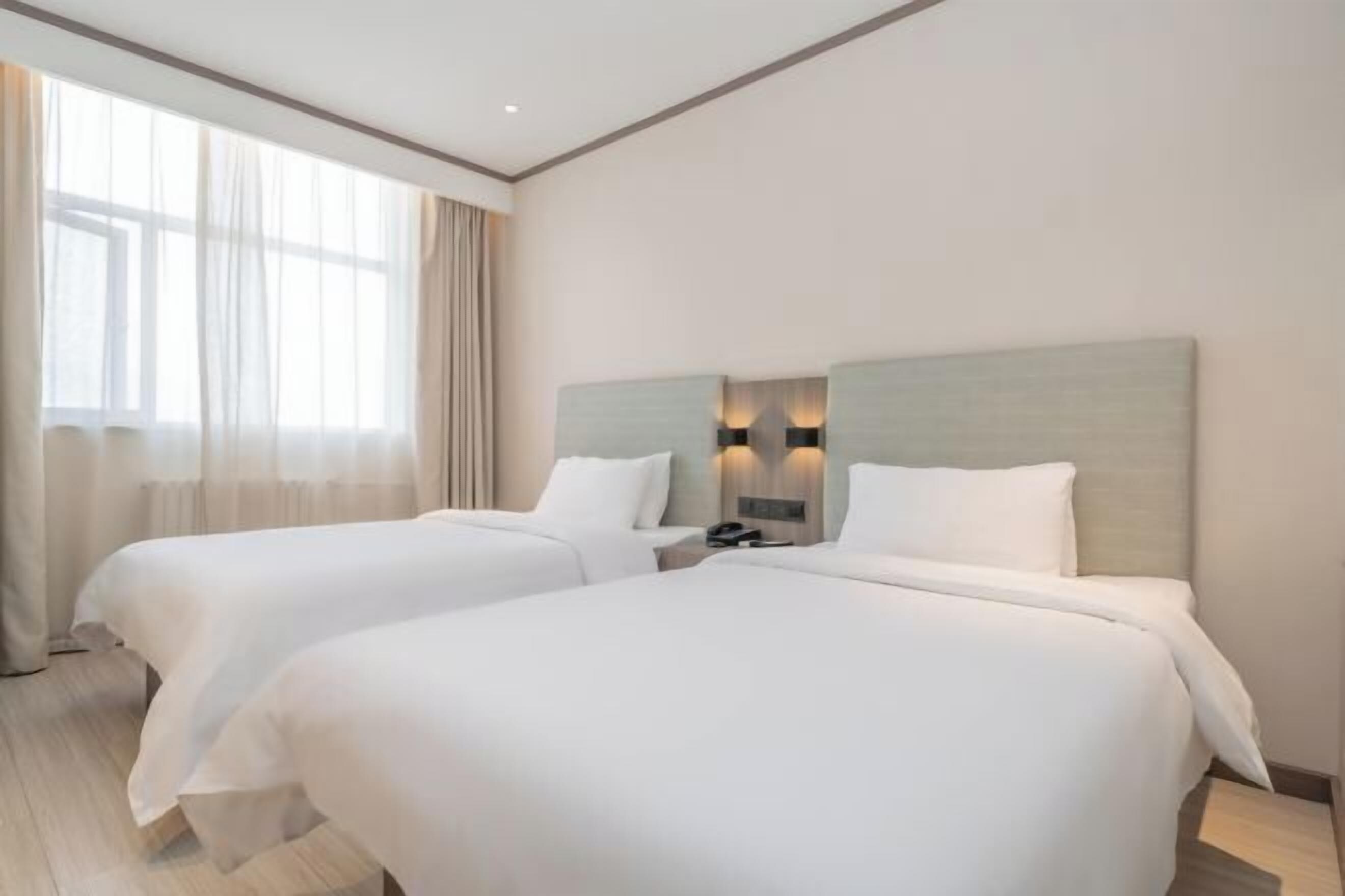 Ein Hotelzimmer mit zwei Betten, einem Nachttisch, einer Lampe und einem großen Fenster mit Vorhängen.