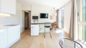 Eine moderne Küche mit weißen Schränken, einem kleinen Kühlschrank, einer Mikrowelle und einem Flachbildfernseher auf einem Regal. Im Essbereich stehen zwei Stühle und ein Tisch.