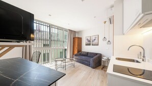 Eine moderne Wohnung mit Küche, Essbereich und Wohnzimmer. Sie verfügt über ein großes Fenster, einen Flachbildfernseher, eine Couch und einen kleinen Tisch.