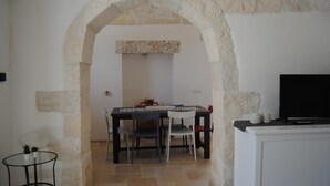 A dining area with a stone archway, a table with chairs, a TV on a cabinet, and a small table with decorative items. - Trullo Sogni Di Pietra - Ostuni (Ostuni)