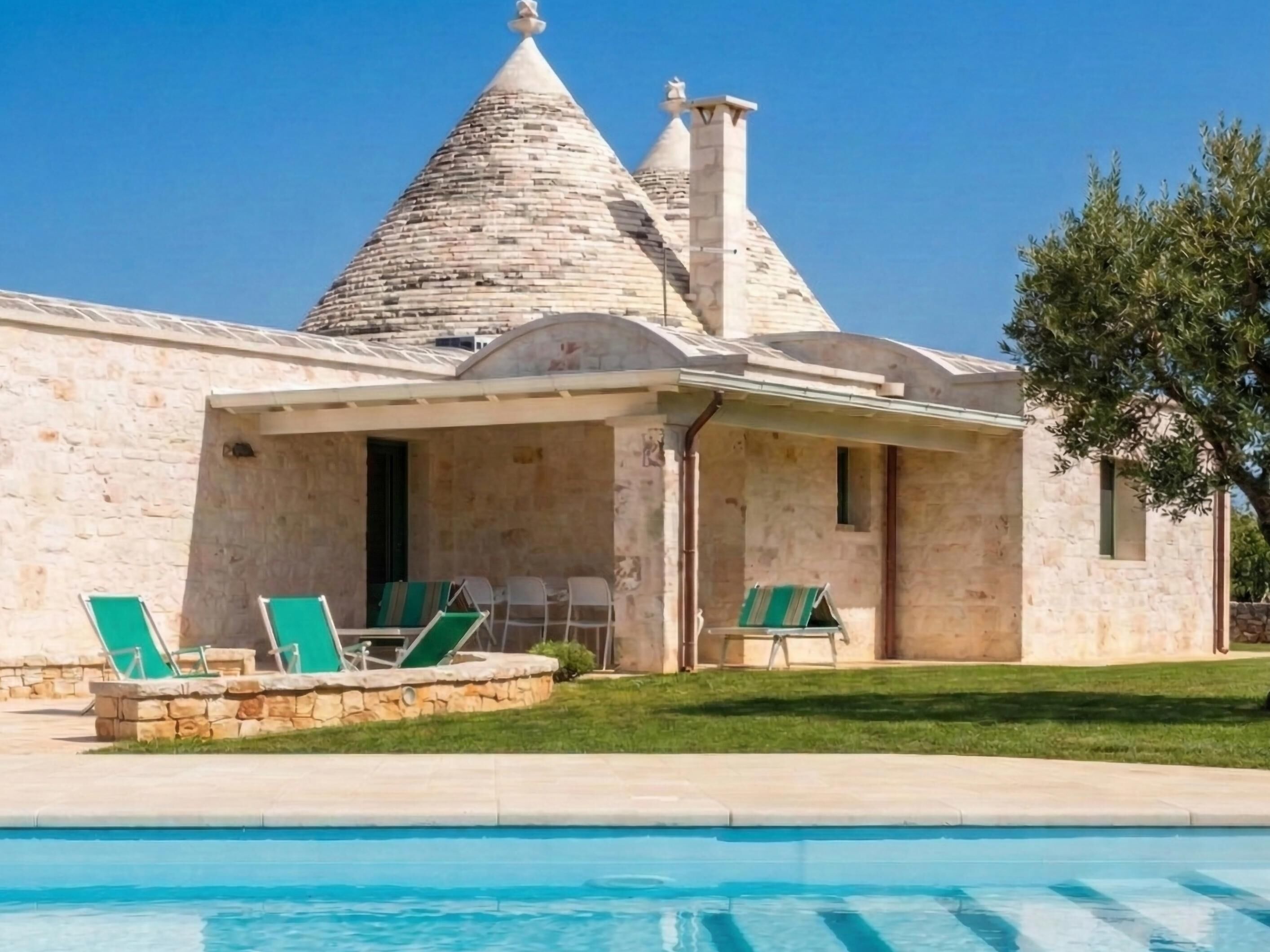 Un edificio de piedra con techo cónico, una piscina y sillones reclinables verdes.