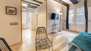 Una habitación moderna con una mesa de comedor de cristal, una silla negra, una cama turquesa, un televisor y una lámpara de pie.