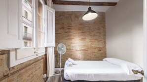 A small, single-bed room with a brick wall, a standing fan, and a hanging light fixture.