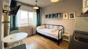 Ett kompakt hotellrum med en enkel säng, ett litet pentry, en matplats och ett fönster med gardiner.