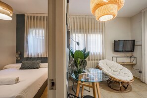 Hotelkamer met een bed, een glazen tafel met stoelen, een kamerplant, een rieten stoel en een tv op een standaard.