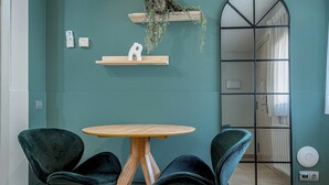 A room with teal walls, a round wooden table, two dark green chairs, a wooden shelf with a plant, a white abstract sculpture, and a large mirror reflecting a bathroom. - Bebarceloner Sants Estudios A5 (Barcelona)