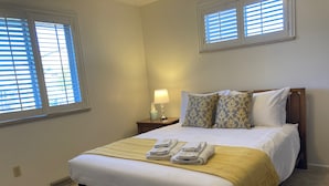 A neatly made bed with a yellow throw blanket, two pillows, and folded towels. There is a bedside table with a lamp and a tissue box. The room has two windows with blinds, a carpeted floor, and a white wall. - Charming, vintage 3-bedroom house in Arvada with AC (Arvada)