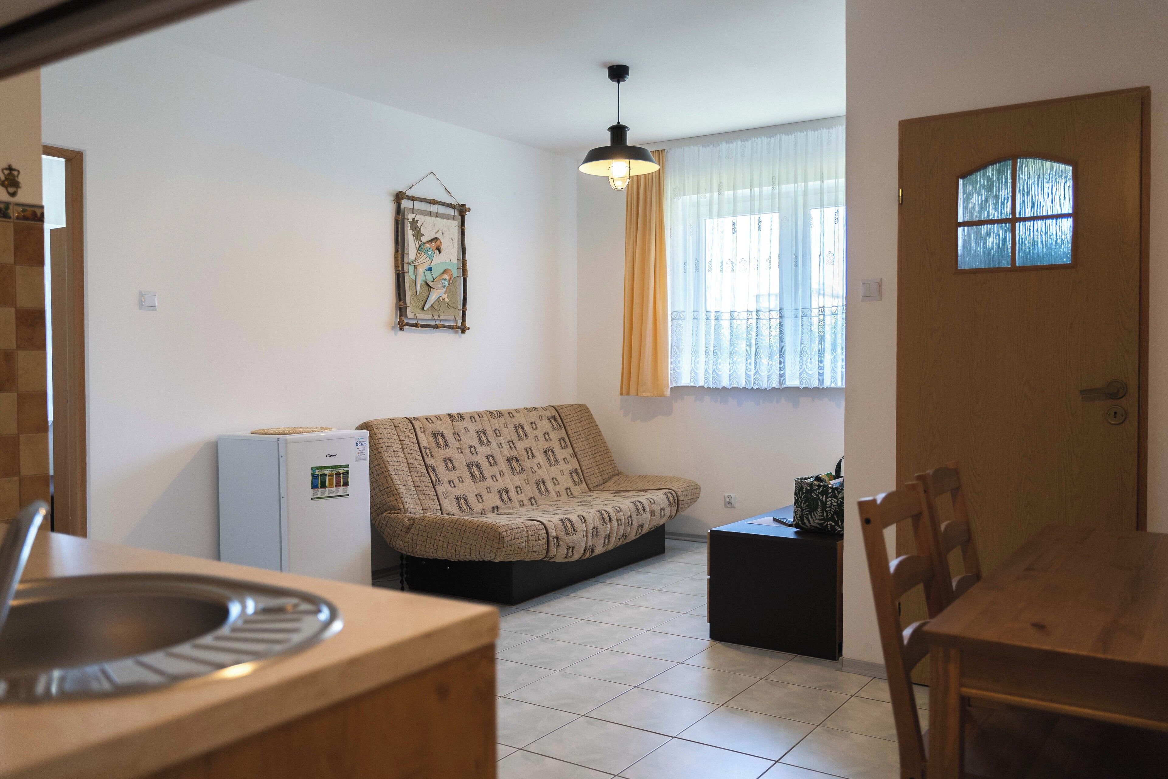 Een ruimtelijk ingerichte leefruimte met een kitchenette, een bank, een eettafel en een raam met gordijnen.