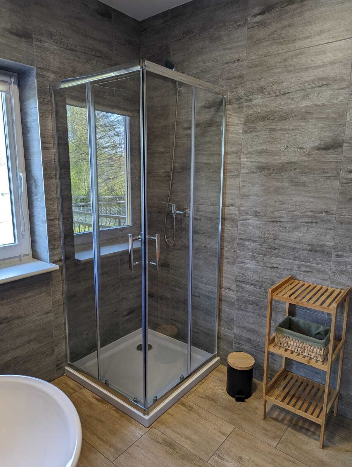 A modern bathroom with a glass-enclosed shower, a wooden floor, and a small wooden shelf with a trash bin.