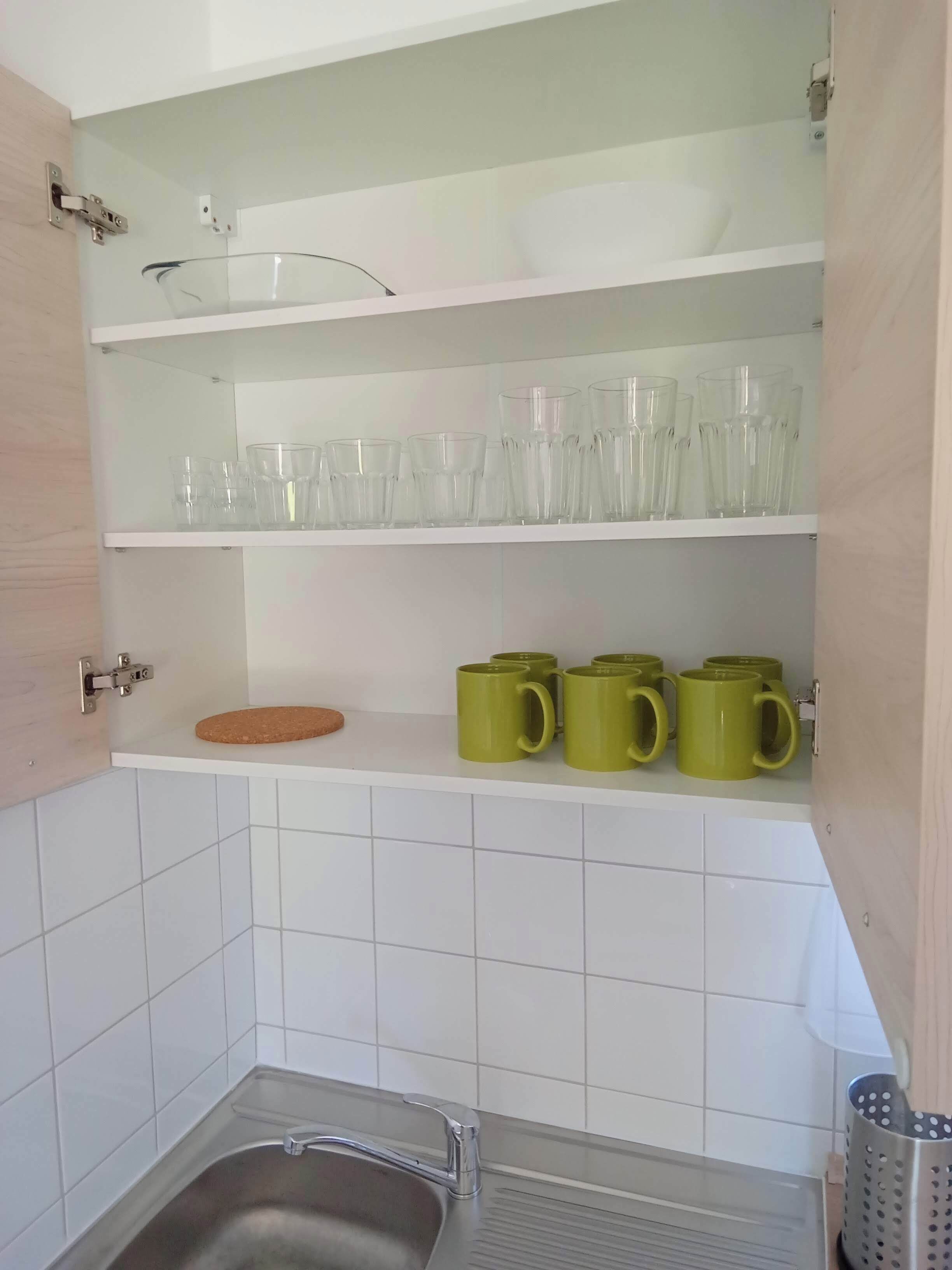 A kitchen with white tiled walls, a stainless steel sink, and shelves holding clear glasses and green mugs.