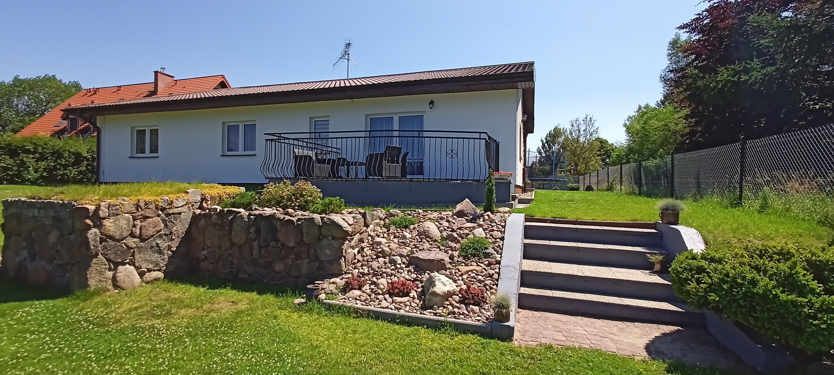 A two-story house with a balcony, stone wall, and a paved pathway leading to the entrance.