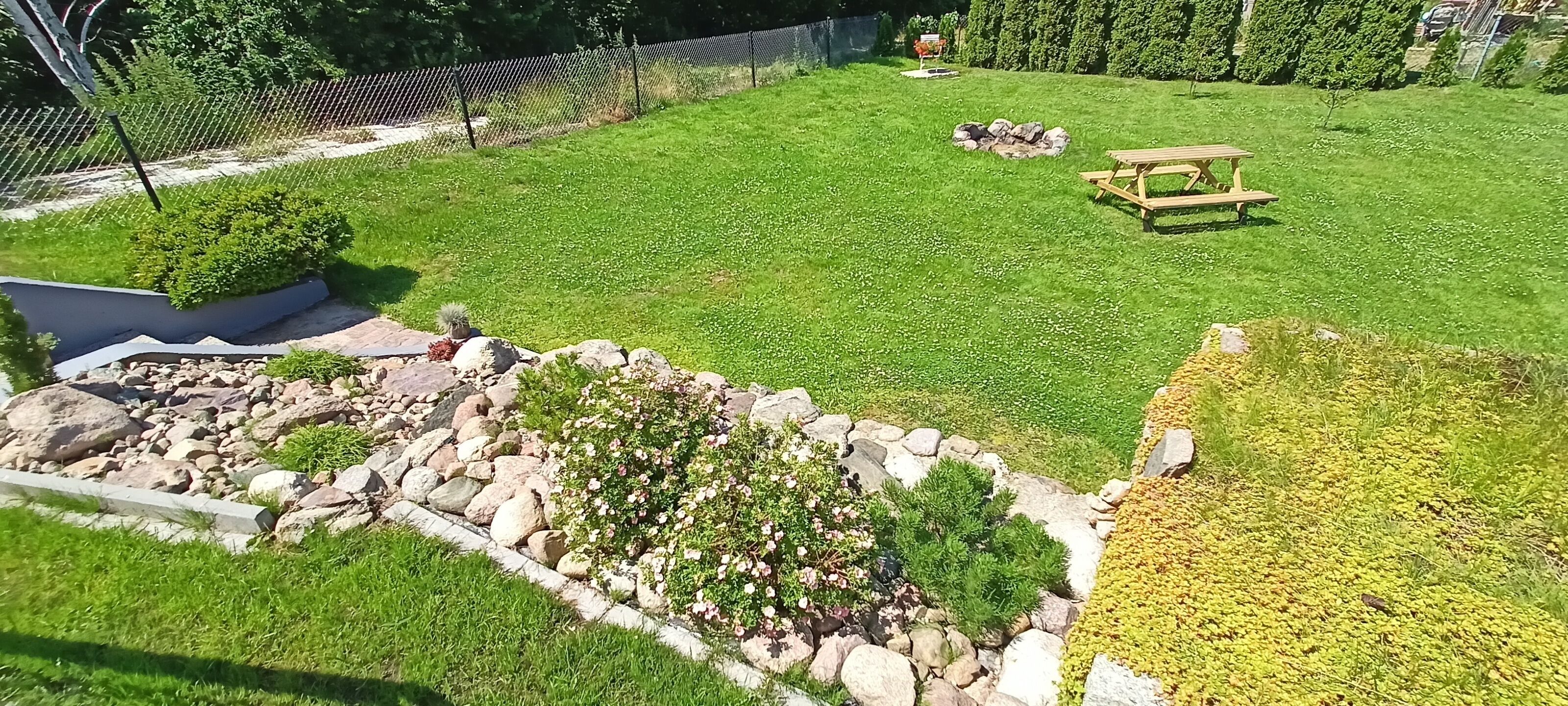 Ein gepflegter Rasen mit einer hölzernen Bank, ein kleiner Gartenbereich mit Steinen und Pflanzen, und ein Zaun im Hintergrund.