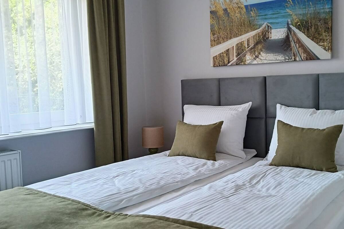 A neatly made bed with white linens and two pillows, a headboard, and a painting of a beach scene on the wall.