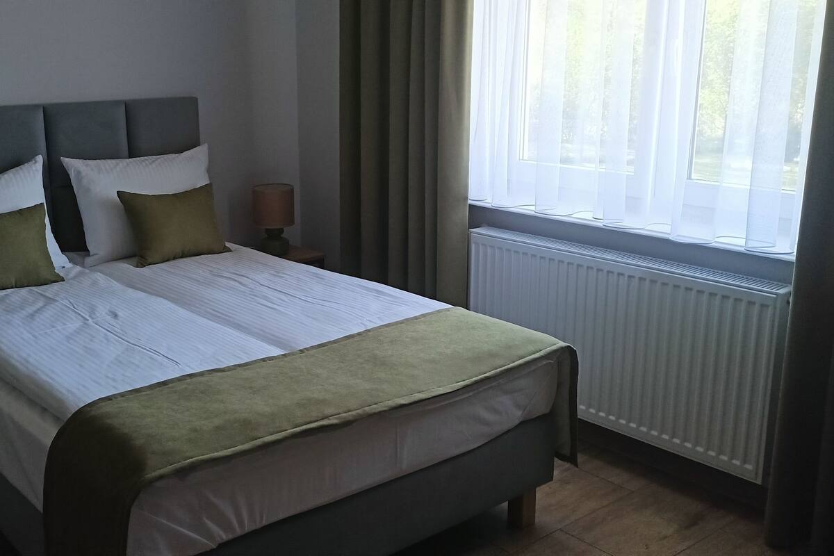 A neatly made bed with a grey headboard, white linens, and green accents. There are two bedside lamps on wooden nightstands. A window with sheer curtains allows natural light into the room.