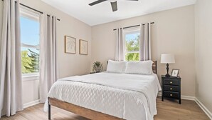A bedroom with a wooden bed, a nightstand, a ceiling fan, and large windows with curtains. - Copper Hill Home in Walnut Creek (Sugarcreek)