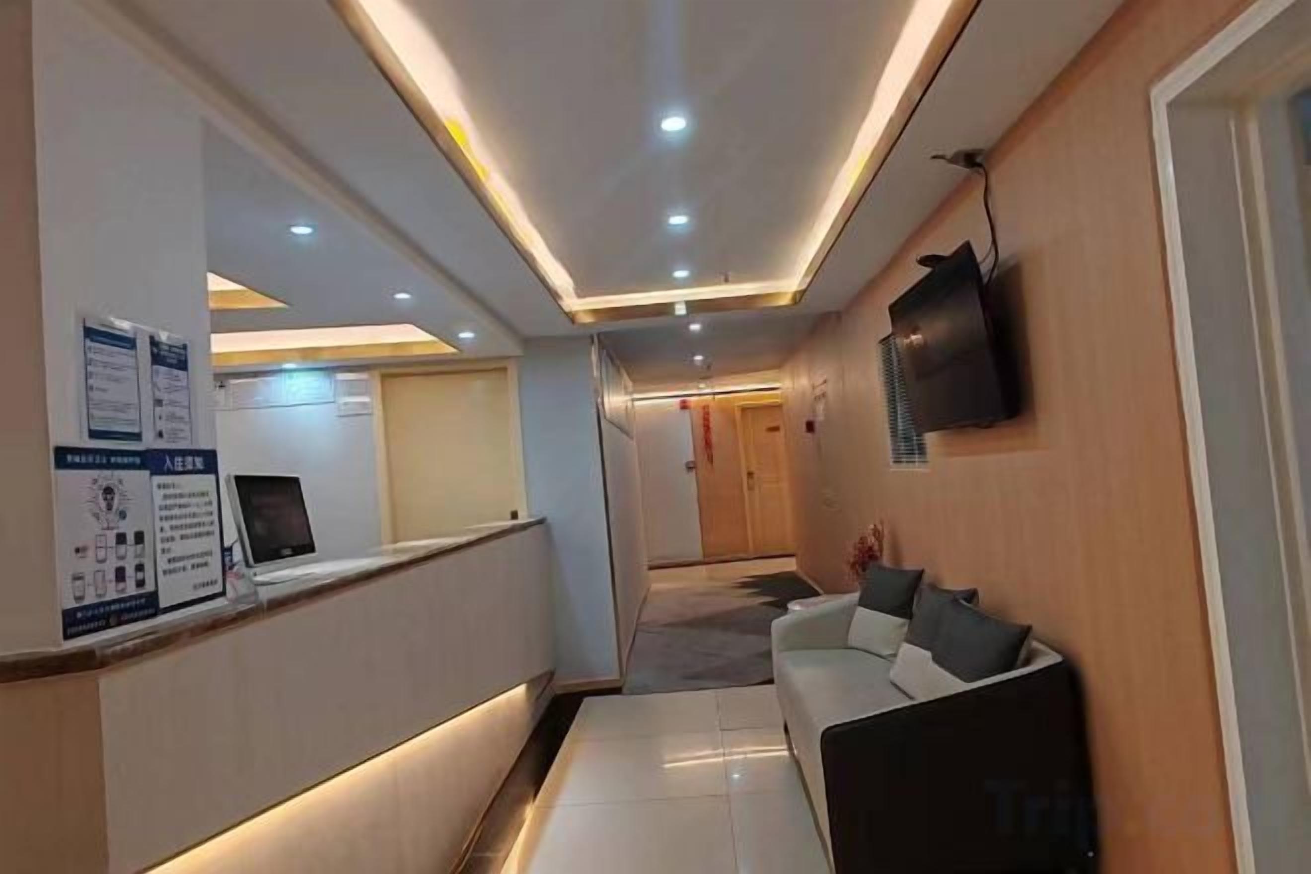 A hotel reception area with a reception desk, a waiting area with a sofa, a TV mounted on the wall, and a corridor leading to other rooms.