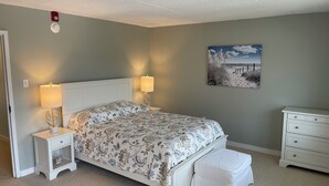 Ein Schlafzimmer mit Bett, Nachttischen, einer Bank, einer Kommode und einem Strandbild an der Wand.