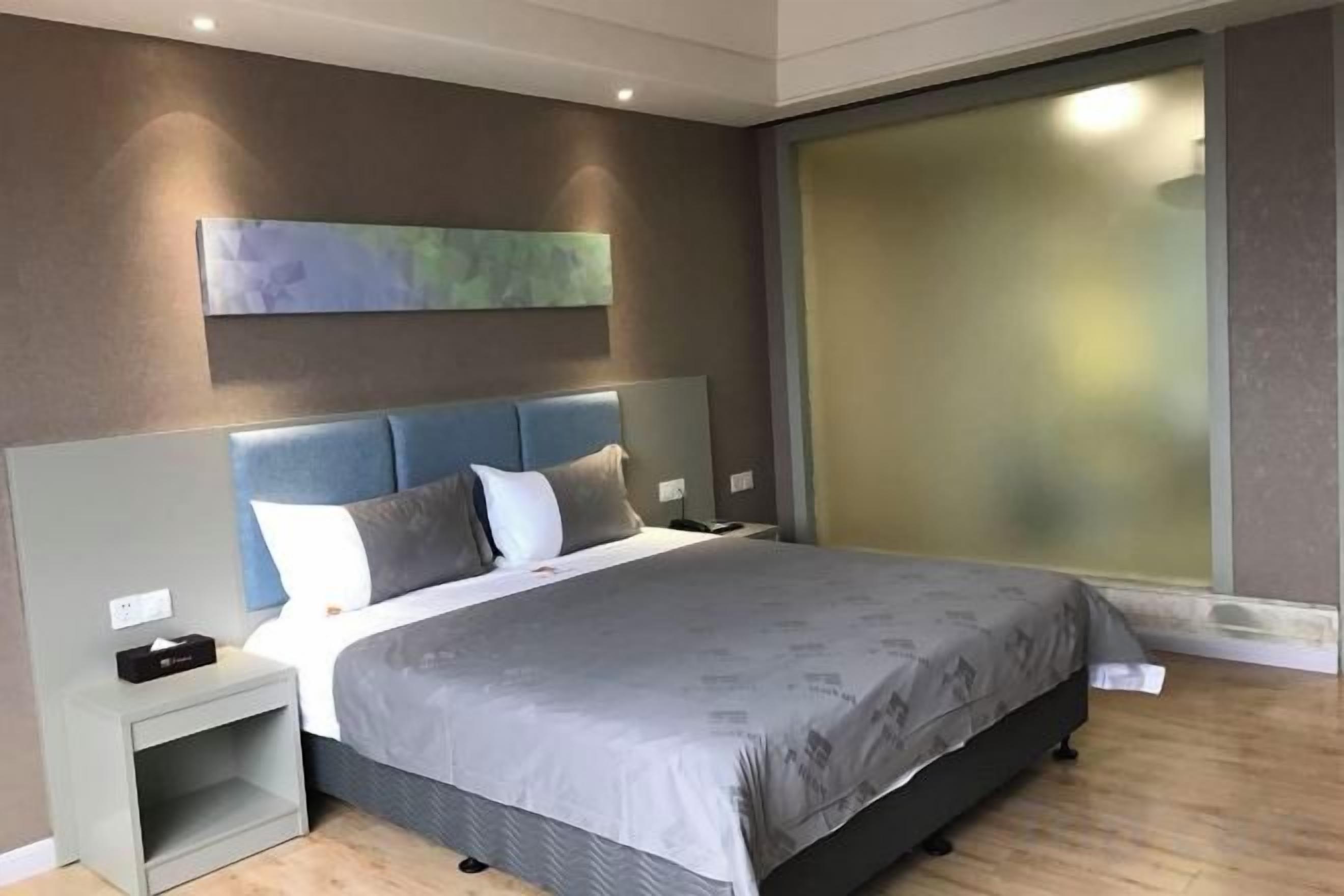 A modern bedroom with a large bed, a nightstand, a wall-mounted artwork, and a frosted glass window.