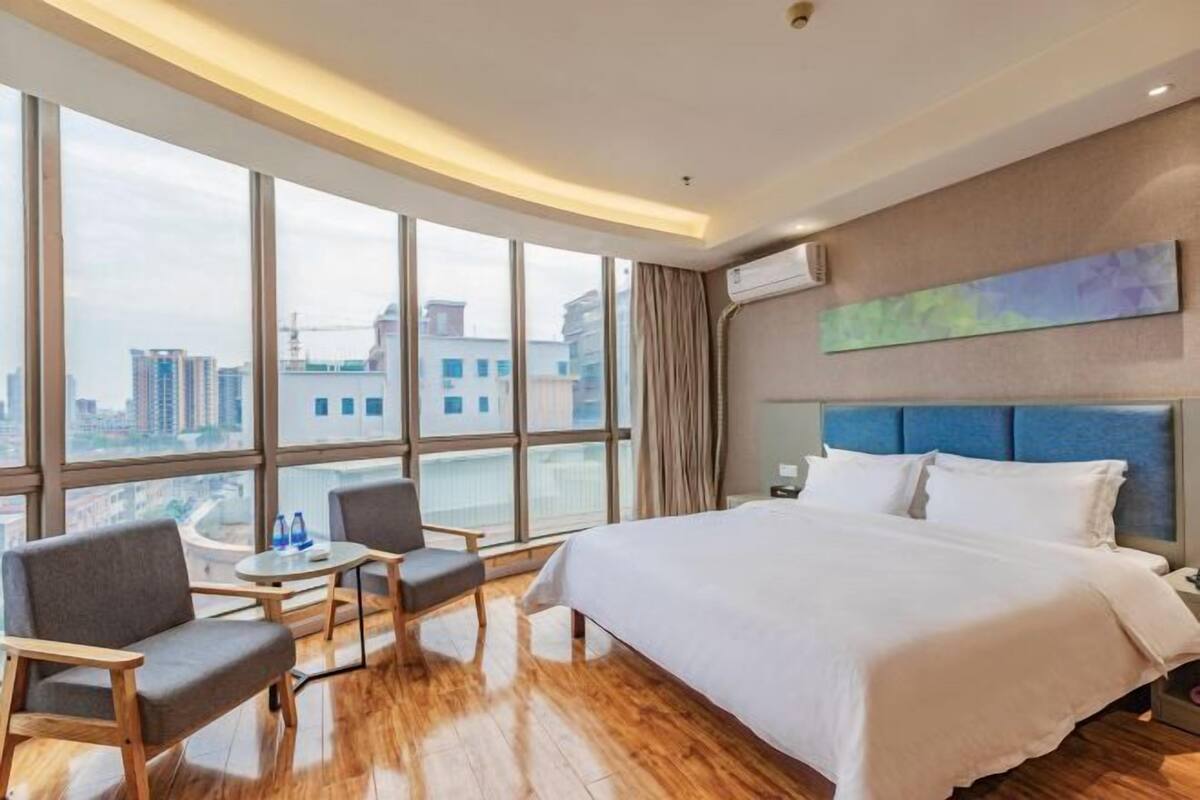 Habitación de hotel con una cama grande, dos sillones, una mesita y vistas a la ciudad a través de amplios ventanales.