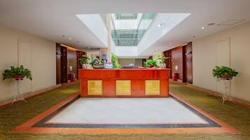 A hotel lobby with a reception desk, potted plants, and a high ceiling with skylights.