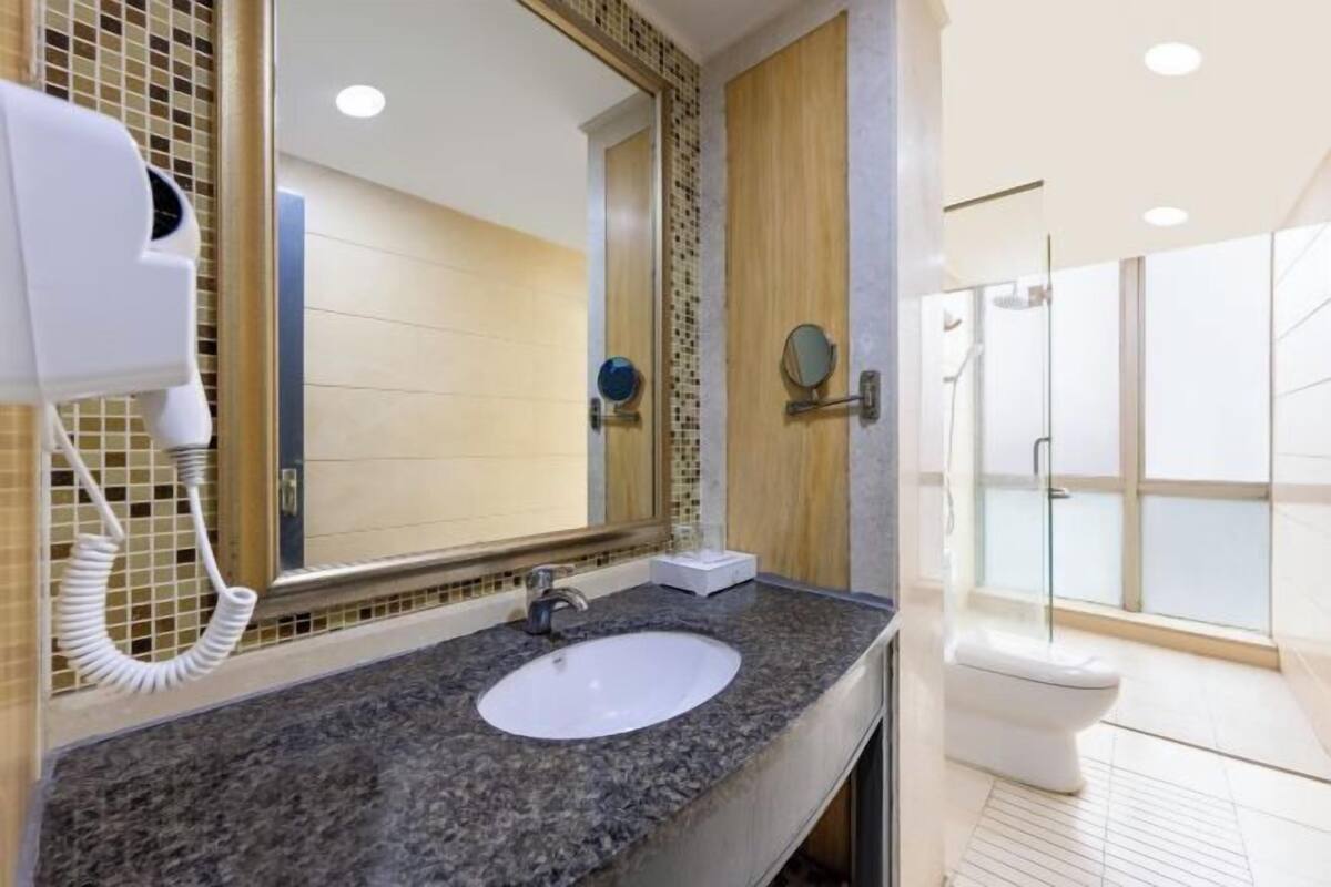 Un baño moderno con encimera de granito, un lavamanos blanco, un espejo grande, un secador de pelo y una ducha con mampara de vidrio.