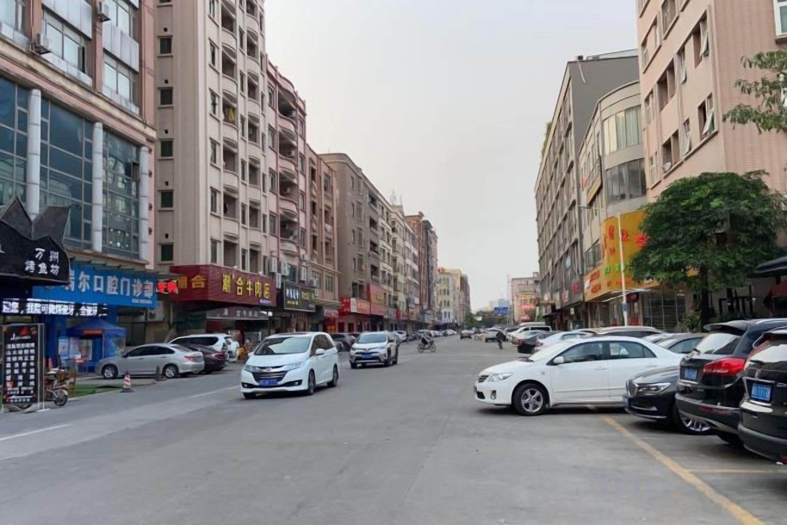 A street lined with multi-story buildings, parked cars, and various storefronts.