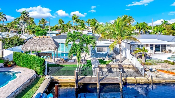 A residential area with a swimming pool, a thatched-roof structure, and multiple houses with large windows. - Pool & Waterfront! Tiki Hut+Close To Beach! (Pompano Beach)