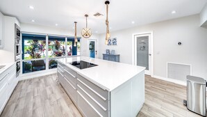 A modern kitchen with a large island, white cabinetry, and a view of a garden through large windows. - Pool & Waterfront! Tiki Hut+Close To Beach! (Pompano Beach)